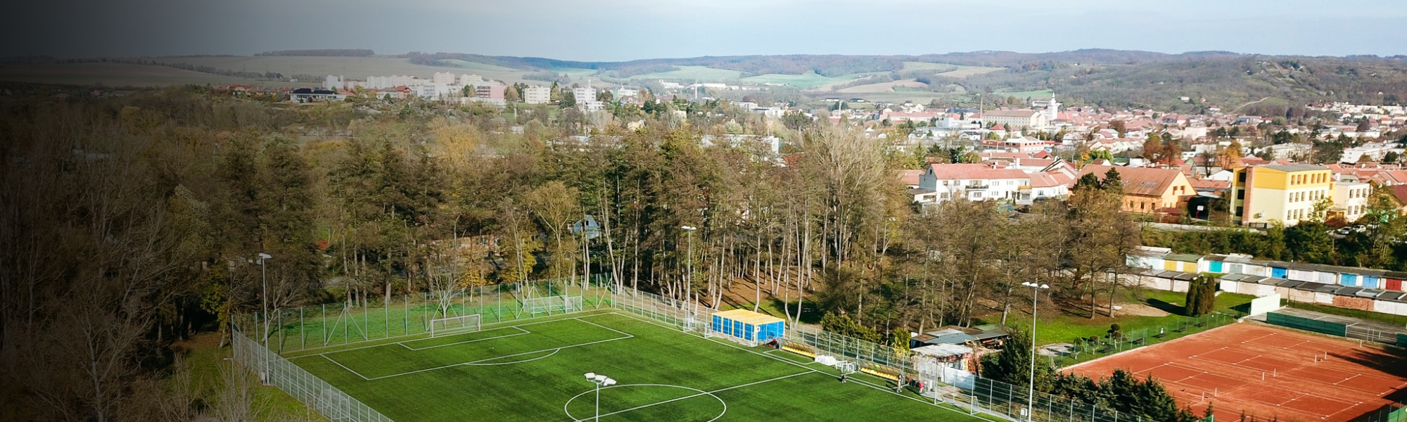 Fußballplatz mit Kunstrasen, gelegen in einer kleinen Stadt, umgeben von Wäldern und Tennisplätzen.