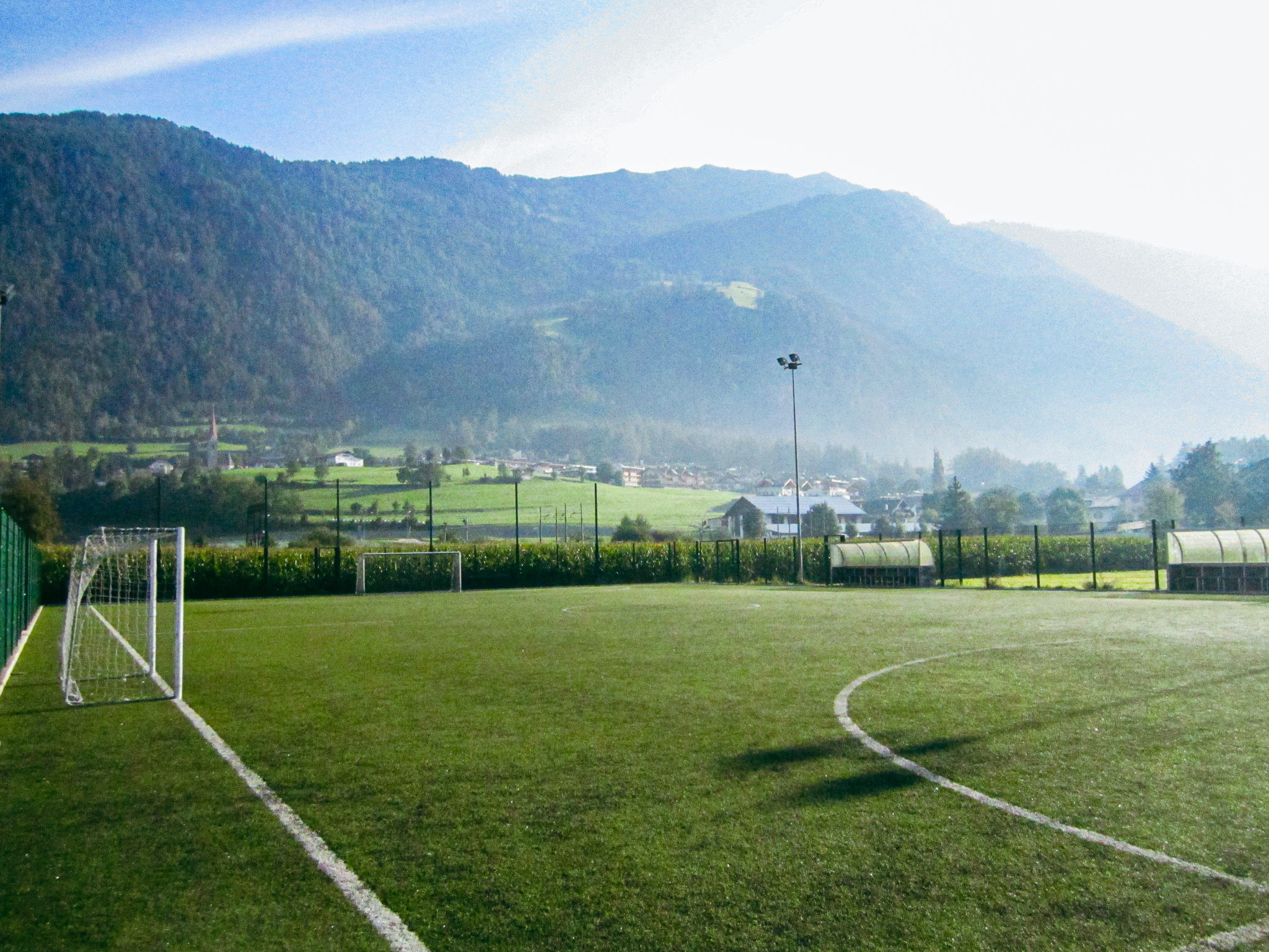 Fußballplatz in Südtirol, eingebettet in eine idyllische Berglandschaft mit Sicht auf ein Tal und umliegende Häuser.