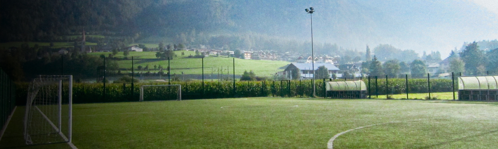 Fußballplatz in Südtirol, umgeben von grünen Feldern, mit Blick auf ein Dorf und Berge im Hintergrund.