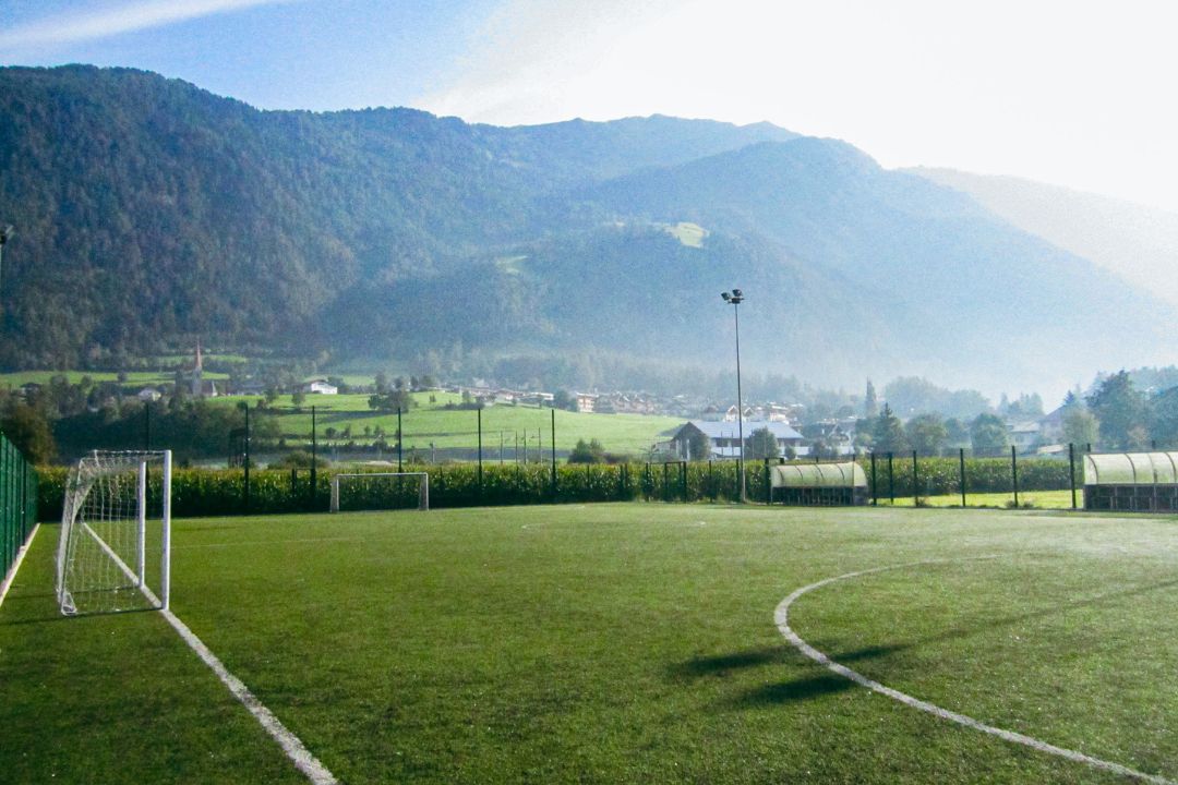 Fußballplatz in Südtirol, eingebettet in eine idyllische Berglandschaft mit Sicht auf ein Tal und umliegende Häuser.