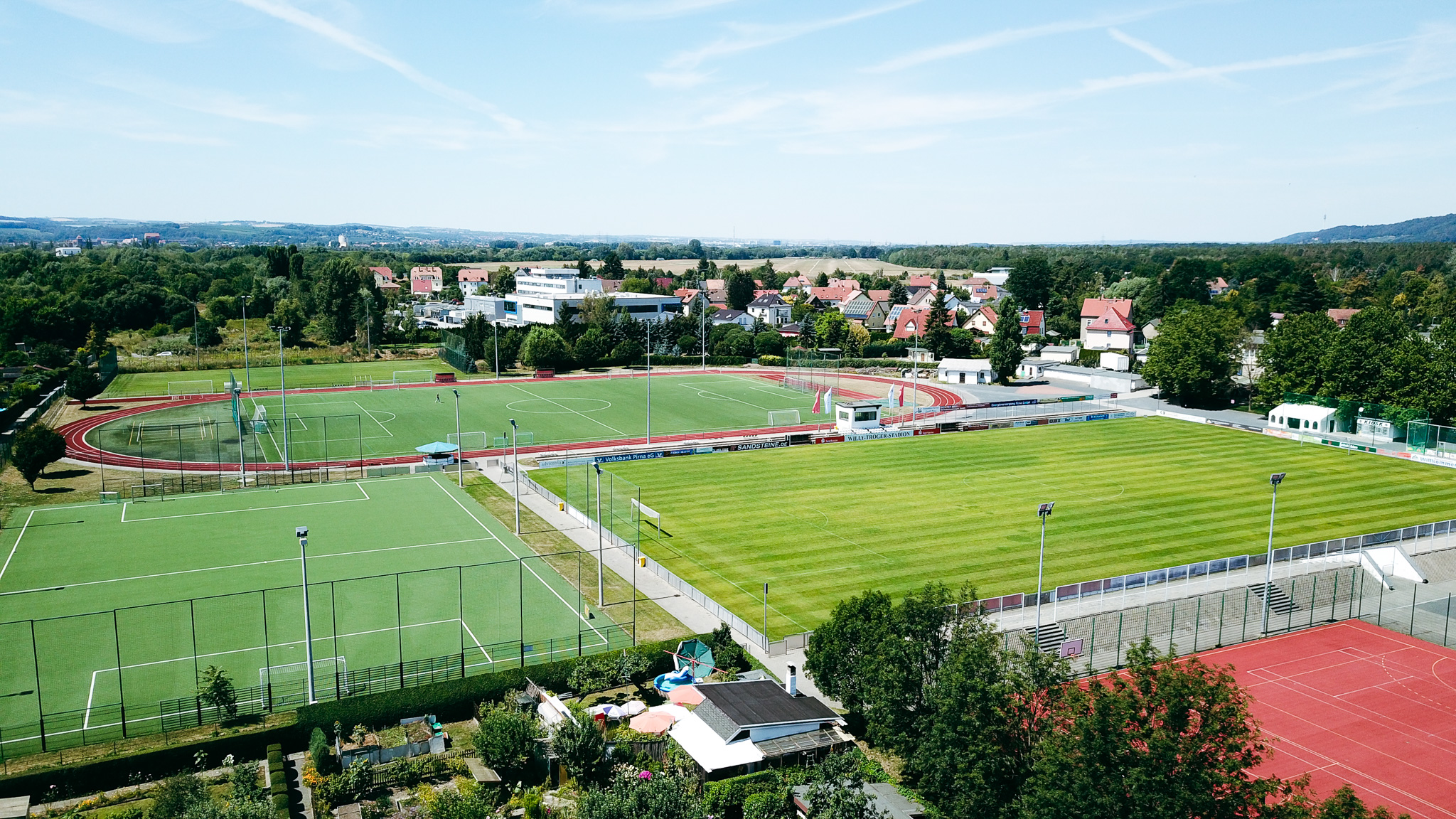 Sportanlage mit Fußballplätzen, Laufbahn und Stadion, umgeben von Wohnhäusern und grüner Landschaft.