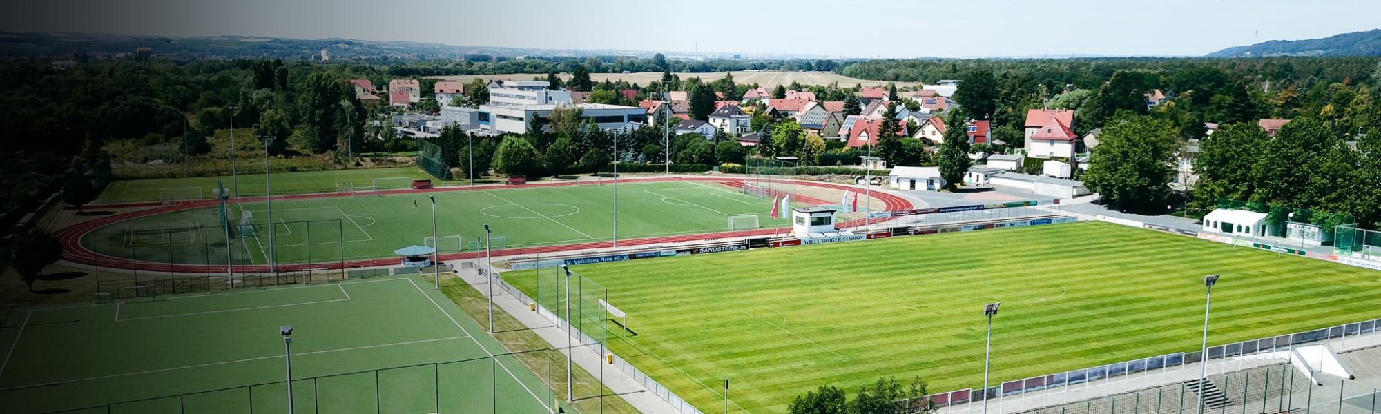 Luftaufnahme von einem Fußballrasenplatz, Kunstrasenplatz und einer Laufbahn, eingebettet in eine Wohnsiedlung und Naturkulisse.