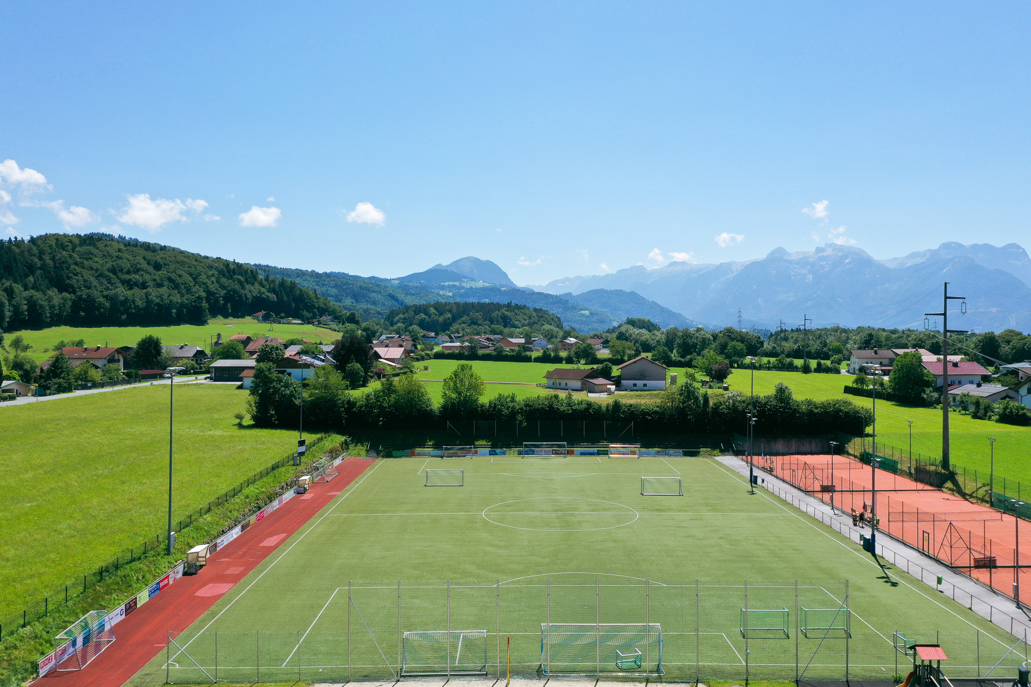 Football pitch in Salzburg, surrounded by green meadows, small houses and picturesque mountains in the background, under a blue sky.