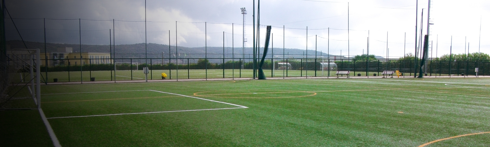 Kunstrasen-Fußballplatz auf Sardinien mit markierten Spielfeldlinien und Umzäunung, im Hintergrund Berge und Gebäude.
