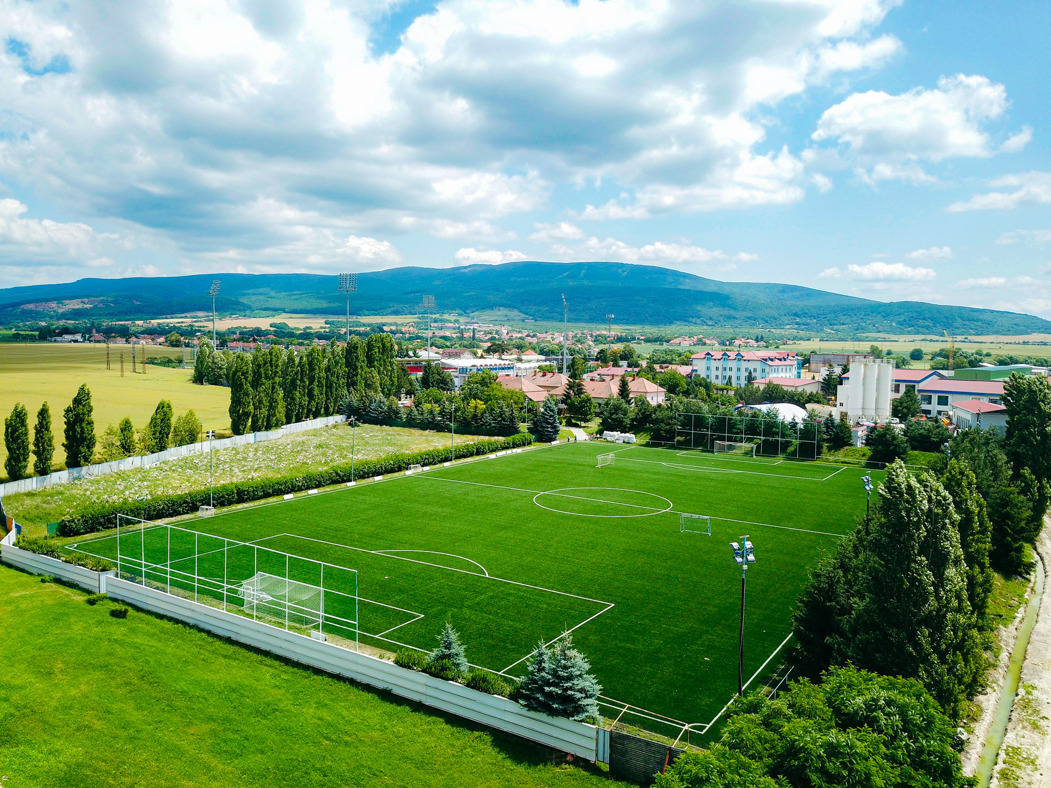 Fußballplatz in der Slowakei, umgeben von grünen Wiesen, Bäumen und Bergen im Hintergrund, mit Flutlichtmasten und Toren.