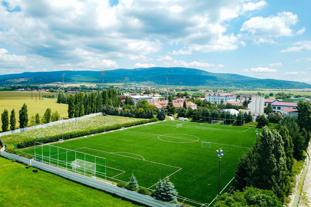 Fußballplatz in der Slowakei, umgeben von grünen Wiesen, Bäumen und Bergen im Hintergrund, mit Flutlichtmasten und Toren.