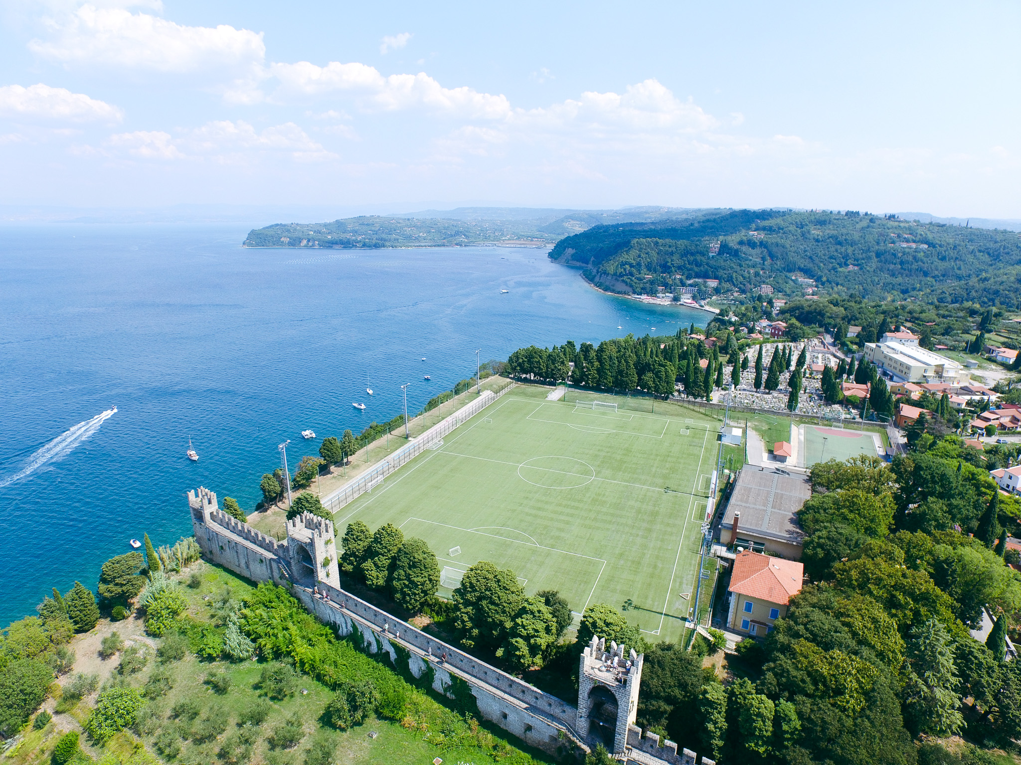 Fußballtrainingsplatz direkt am Meer in Slowenien, mit Blick auf die Küste, die umliegenden Hügel und ein kleines Dorf mit typischen südeuropäischen Gebäuden.