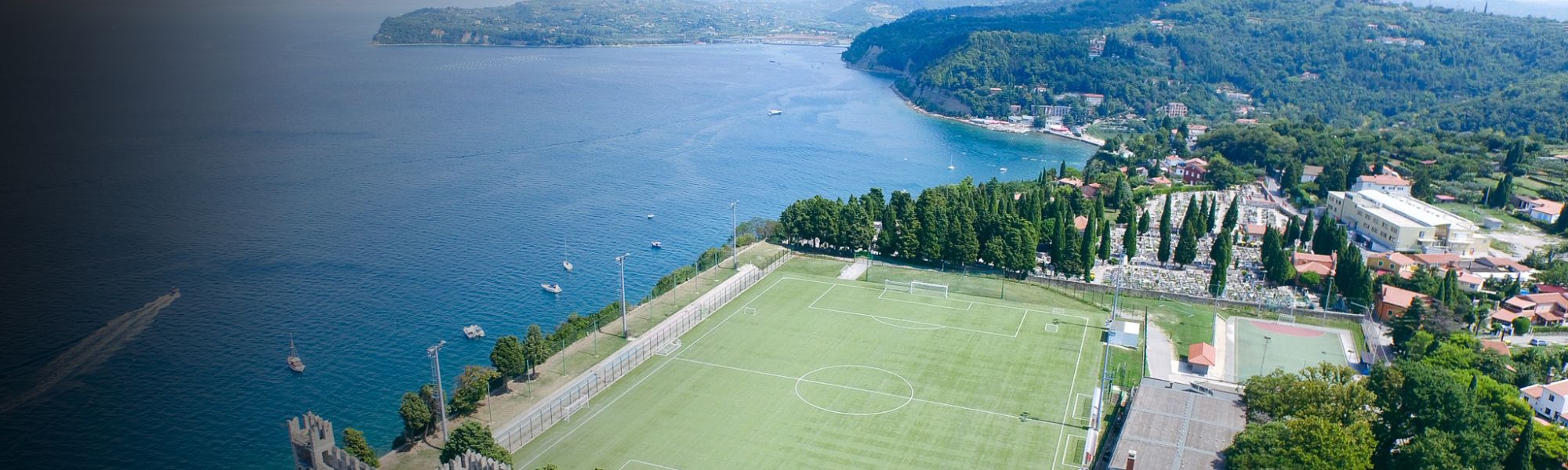 Luftaufnahme eines Fußballplatzes an der Adriaküste in Slowenien, umgeben von grüner Landschaft und kleinen Booten auf dem Wasser.