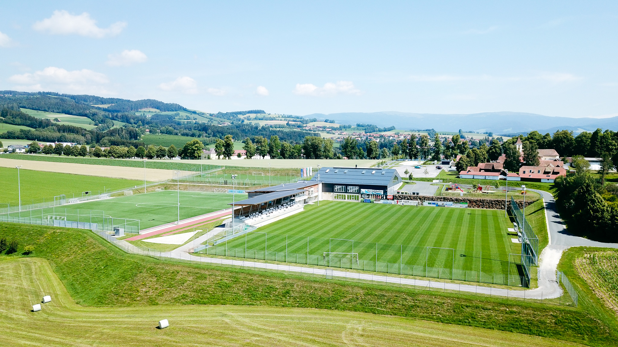Modern football facility in Styria, surrounded by green hills and meadows, with a well-tended natural grass pitch, grandstand and sports building in the background.