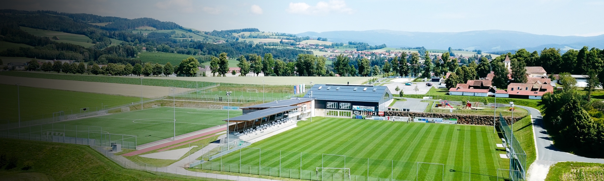 Panoramic shot of a football pitch in Styria, embedded in an idyllic hilly landscape, with grandstand, training ground and club building.
