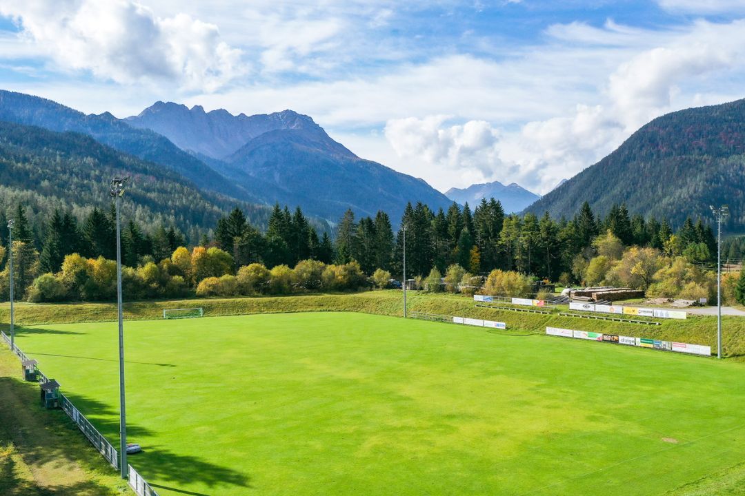 Football pitch in Tyrol, surrounded by mountains and dense forests, with floodlight masts and advertising banners on the edge of the pitch.