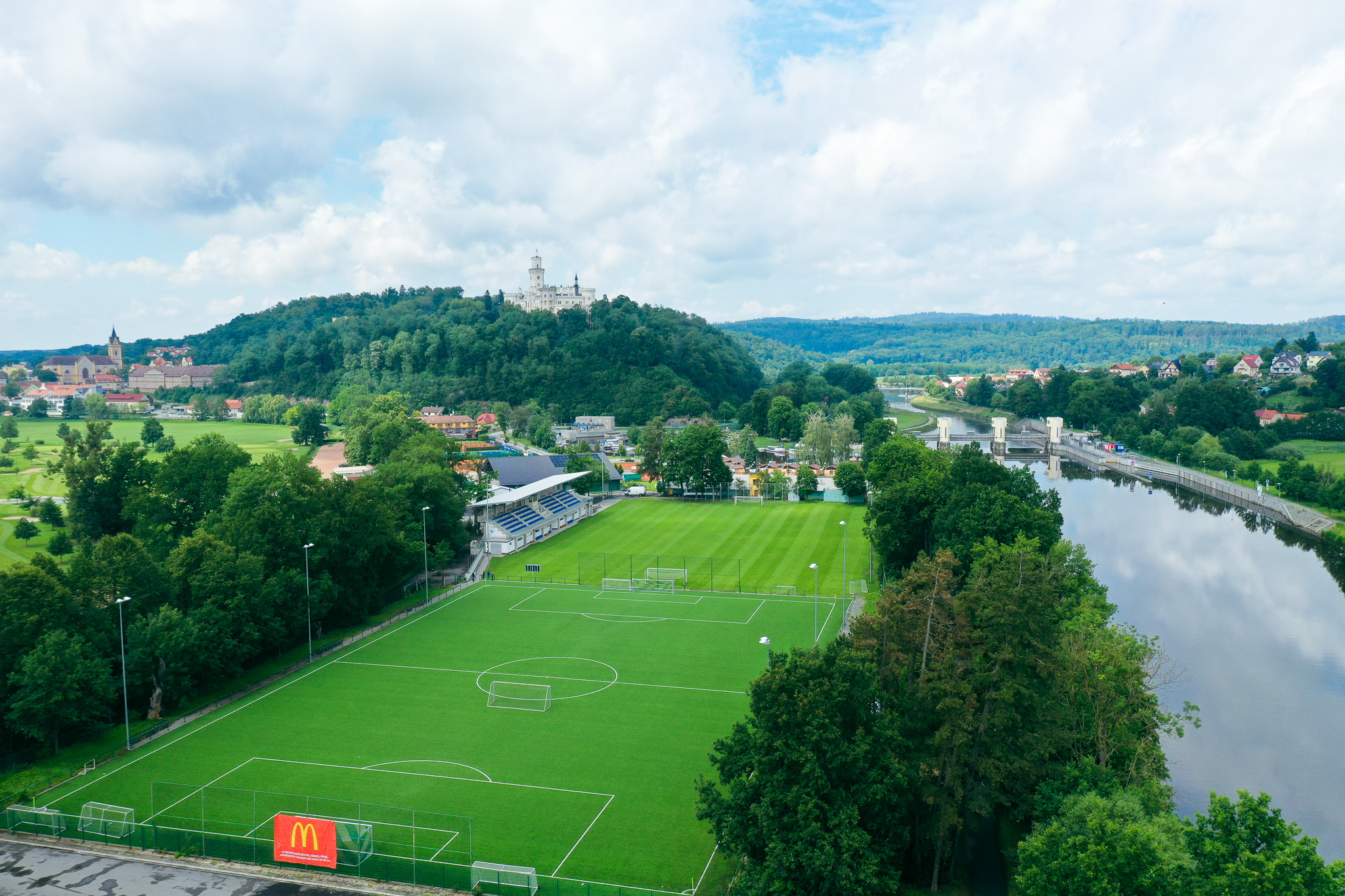Football pitch in the Czech Republic, surrounded by forest and hills, with a view of a castle and a town.