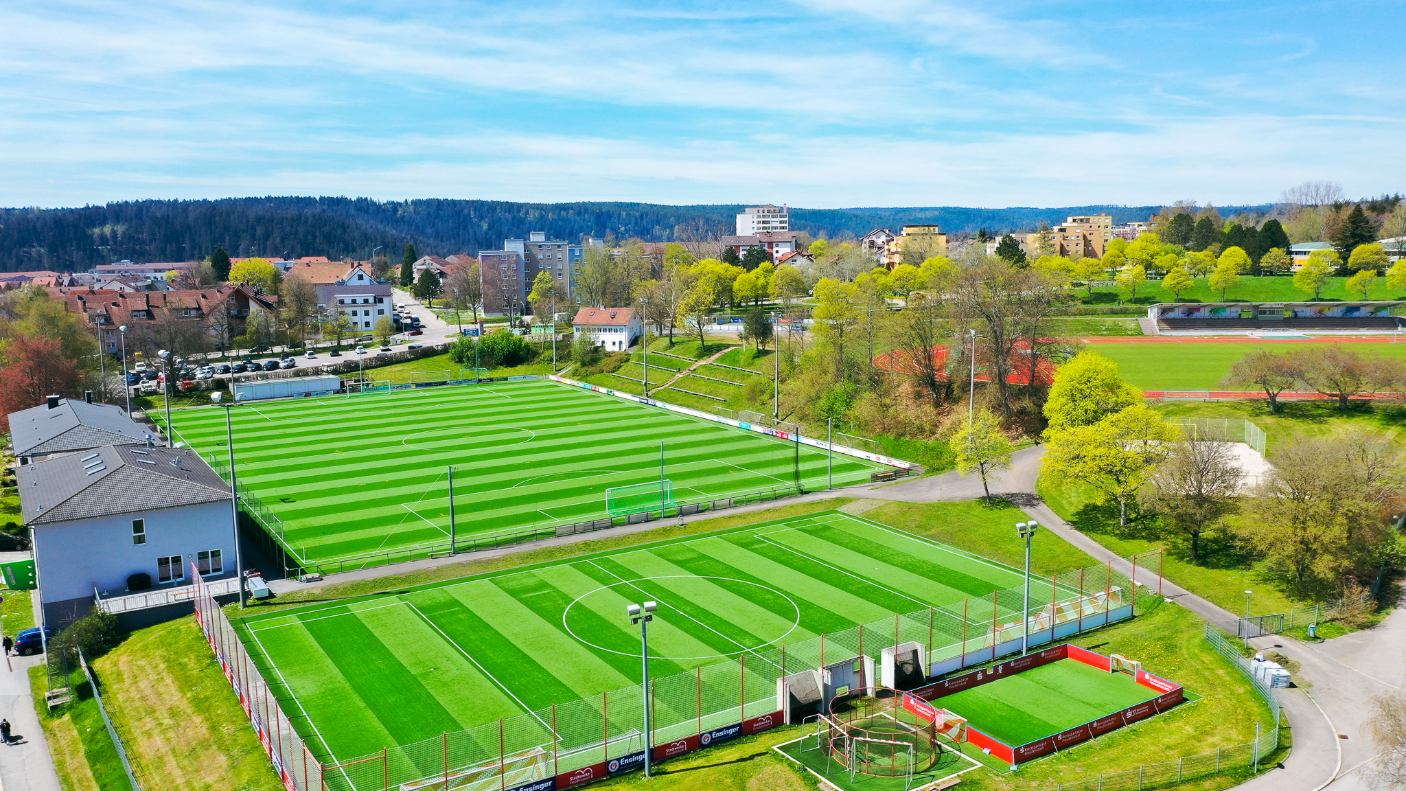 Aerial view of the football training facility in Baden-Württemberg with two artificial turf pitches, small pitch, floodlights and grandstand, embedded in green surroundings.
