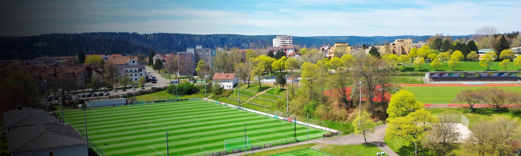 Aerial view of a modern sports facility in Baden-Württemberg with several artificial turf pitches, an athletics facility and surrounded by trees and residential buildings.