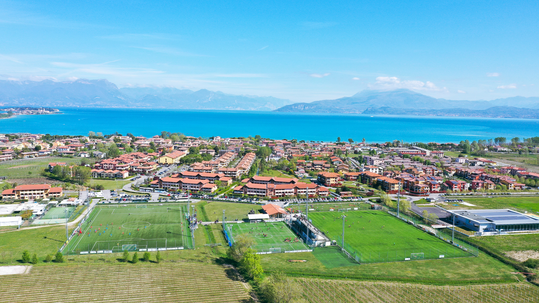 Aerial view of a football training facility on Lake Garda, surrounded by red residential buildings, with the blue lake and mountains in the background.
