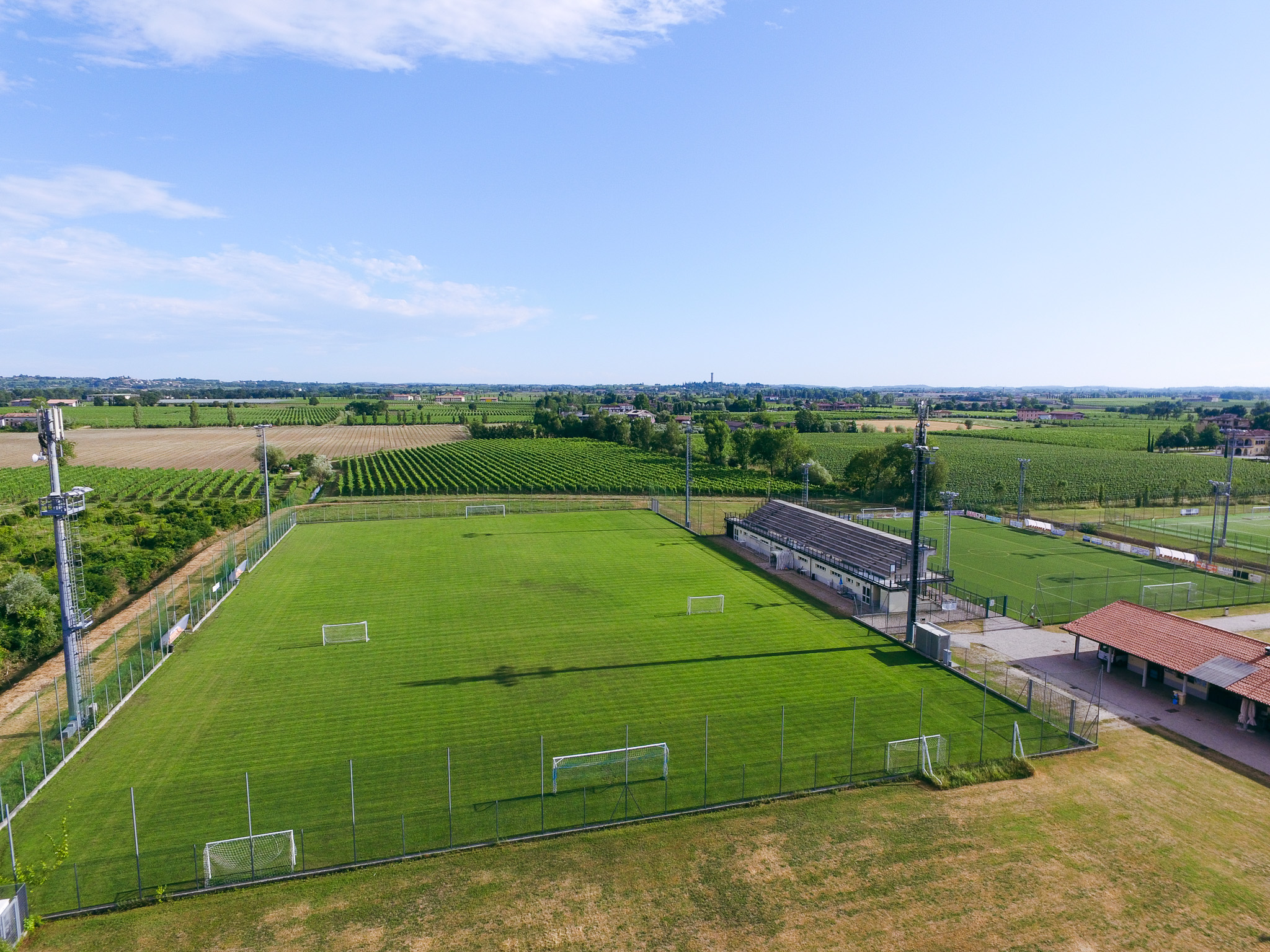 Aerial view of a sports facility with several football pitches, surrounded by a small Italian town on the shore of a lake, with mountains in the background.