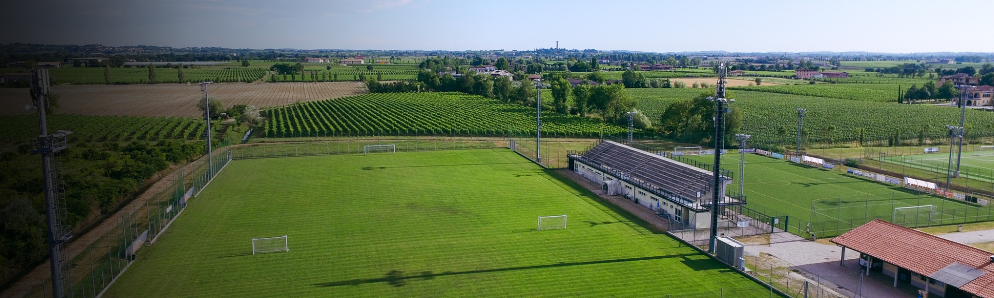 Panoramic aerial view of a sports facility with several football pitches, surrounded by a small Italian town on the shore of a lake.