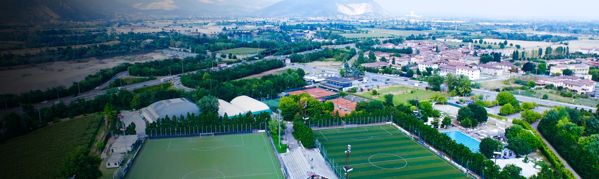 Aerial view of a sports facility in Lombardy with several football pitches, tennis courts, a grandstand and an indoor swimming pool, surrounded by agricultural fields and a small village.
