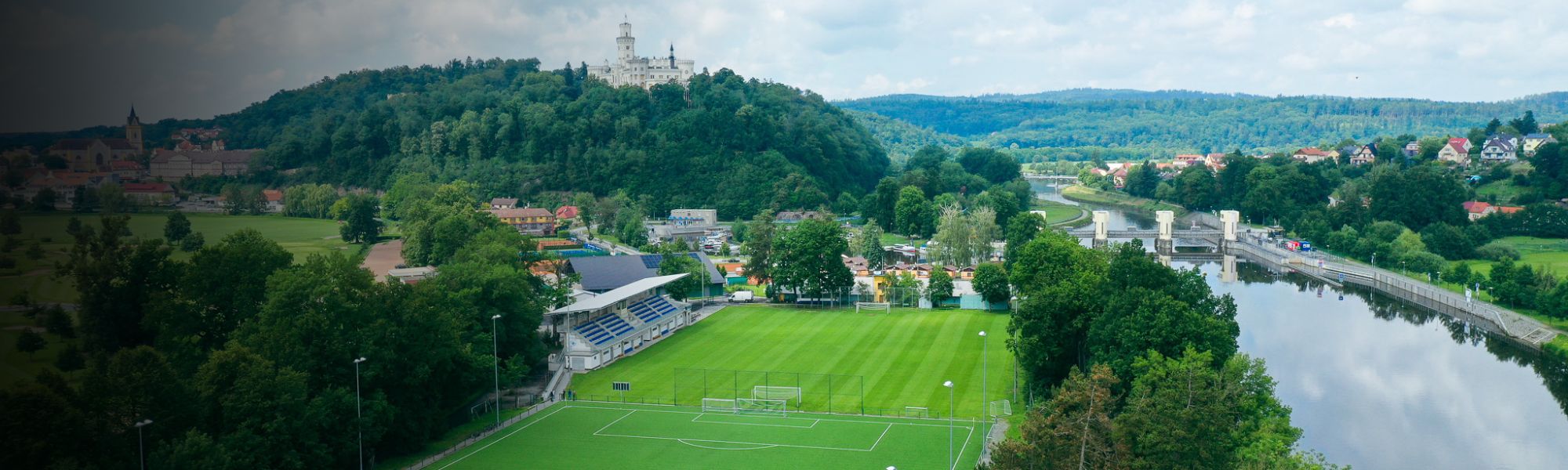 Aerial view of a football pitch in the Czech Republic, next to a river, with a castle and town in the background.