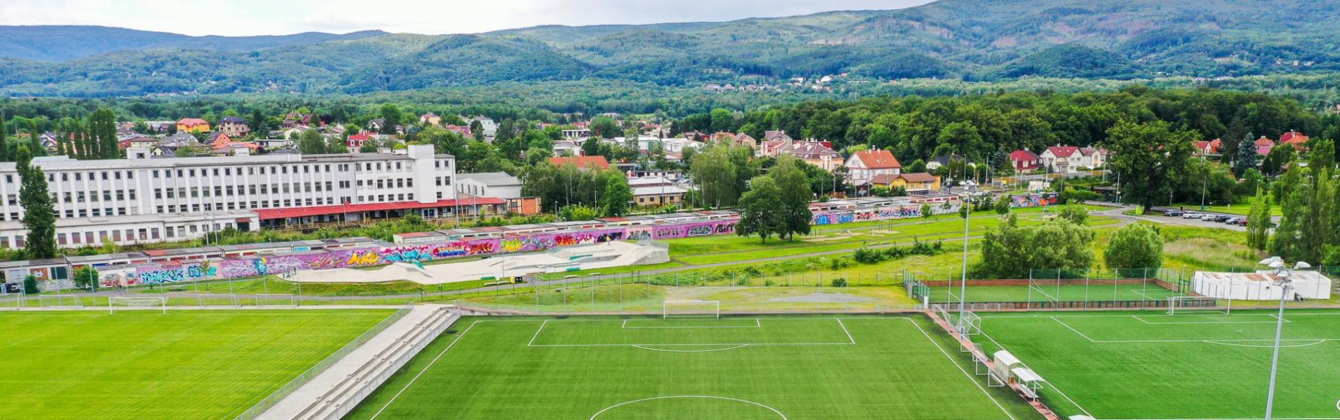  Voetbalveld met kunstgras in een groene omgeving, omringd door heuvels en met een stadsgezicht op de achtergrond.