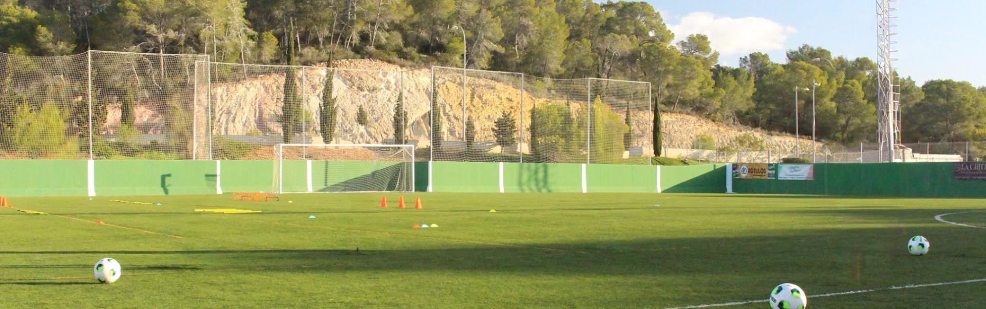 Een voetbalveld met een groene omheining, oranje trainingskegels, ladders, netten achter het doel en bomen en rotsachtige heuvels op de achtergrond onder een heldere hemel.