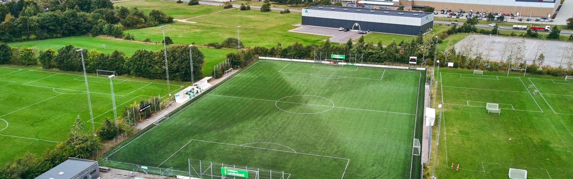 Luchtfoto van een modern voetbaltrainingscomplex in Denemarken met een groot kunstgrasveld, meerdere natuurgrasvelden, verlichtingsinstallaties en aangrenzende industri&euml;le en bedrijfsgebouwen op de achtergrond.
