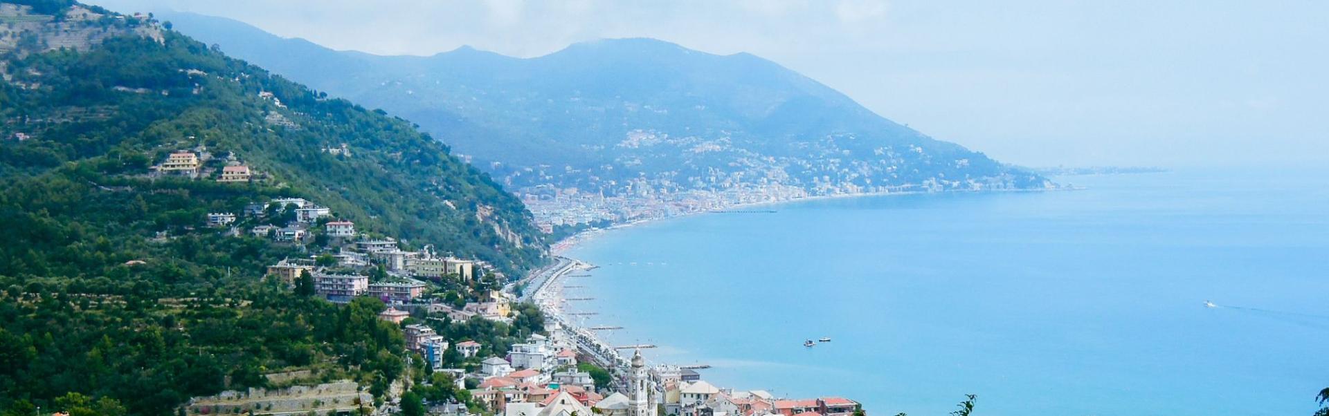 Panoramaansicht der Italienischen Riviera mit der K&uuml;stenstadt, die sich entlang des blauen Meeres erstreckt, umgeben von h&uuml;geliger Landschaft und gr&uuml;ner Vegetation.