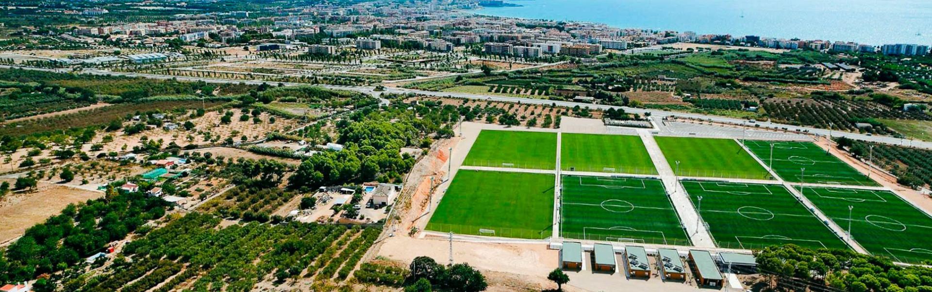 Een moderne sportfaciliteit met meerdere voetbalvelden, uitzicht op de kust en een stedelijke omgeving.