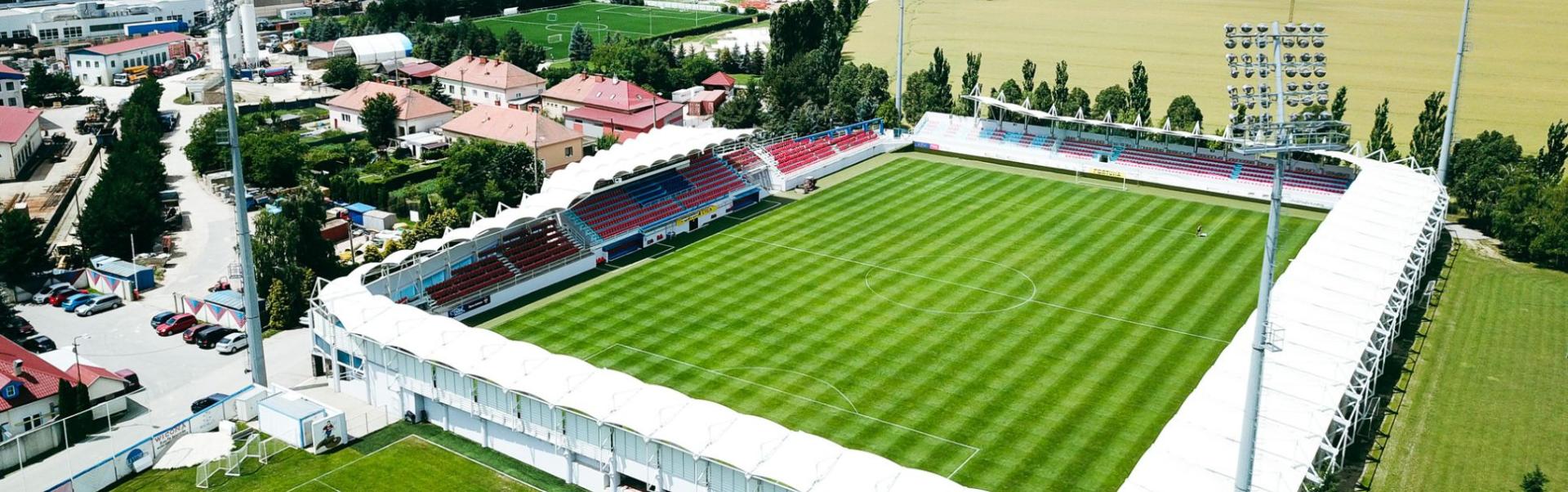 Voetbalstadion in een landelijke omgeving met tribunes, een verlichtingsinstallatie en een weids landschap op de achtergrond.