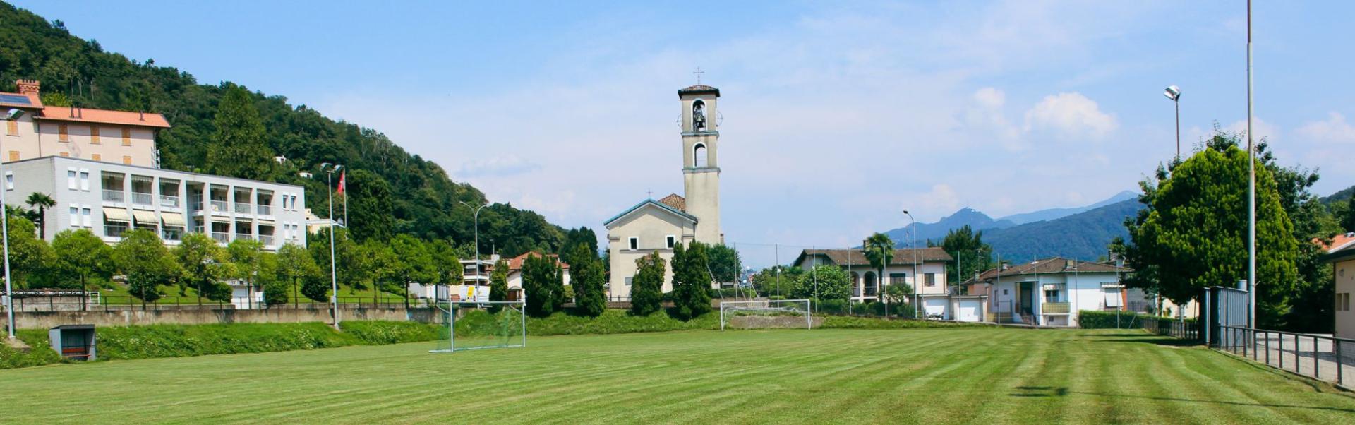 Panoramafoto van een voetbalveld in Ticino, gelegen in een groen landschap met op de achtergrond een kerk en typisch Italiaanse huizen. Perfecte omstandigheden voor een voetbaltrainingskamp in Zwitserland.