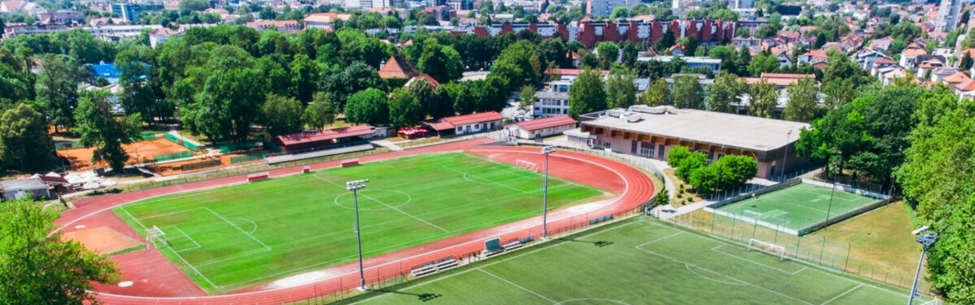 Voetbaltrainingskamp met meerdere velden, een atletiekbaan en tennisbanen, gelegen in een stedelijke omgeving met de bergen op de achtergrond.