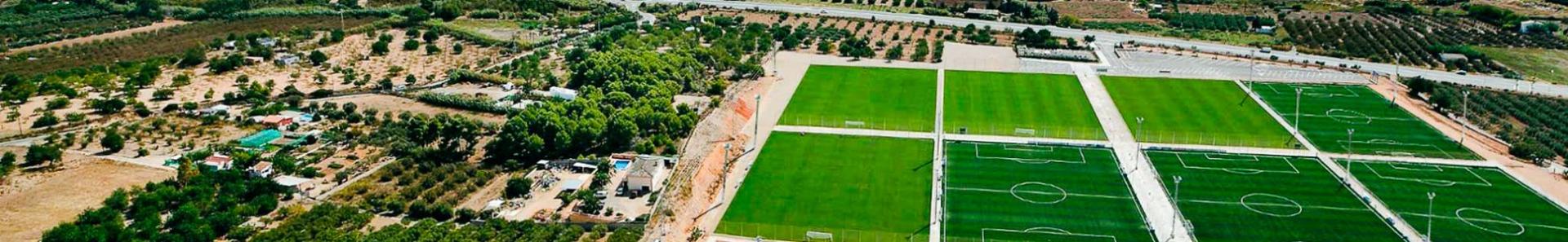 Een moderne sportfaciliteit met meerdere voetbalvelden, uitzicht op de kust en een stedelijke omgeving.