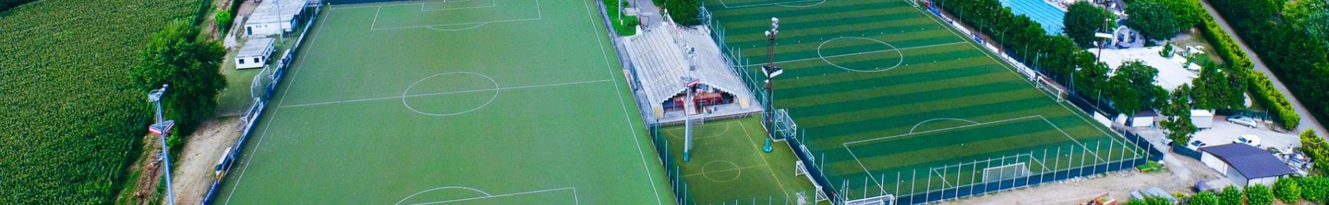Uitgebreide weergave van een trainingsfaciliteit in Lombardije met voetbalvelden, tennisbanen en een overdekt zwembad, ingebed in een groen landschap met bergen en velden op de achtergrond.
