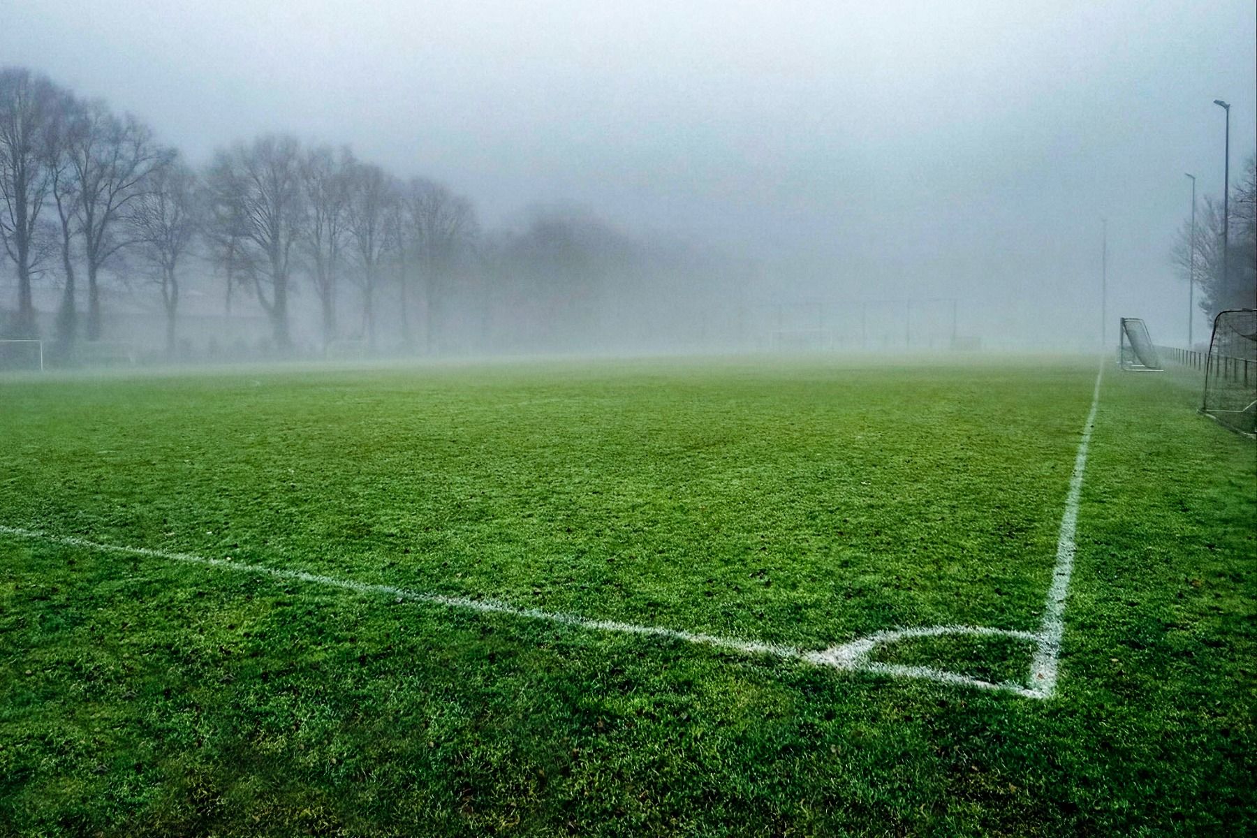 Een voetbalveld met een felgroen veld bij Sporthotel Bruurs in Baarle-Nassau is gehuld in dichte mist, waardoor een koud, mistig tafereel ontstaat - ideaal voor een geconcentreerd trainingskamp bij Sporthotel Bruurs.