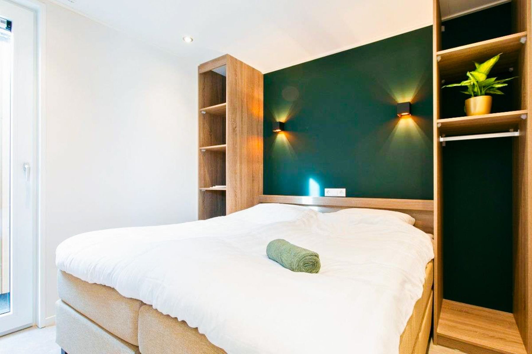 Een moderne, minimale slaapkamer op Duinrell biedt een tweepersoonsbed met wit beddengoed en een groene handdoek, houten planken die een donkergroene accentmuur omlijsten, en natuurlijk licht - ideaal om tot rust te komen na het genieten van sportinfrastr