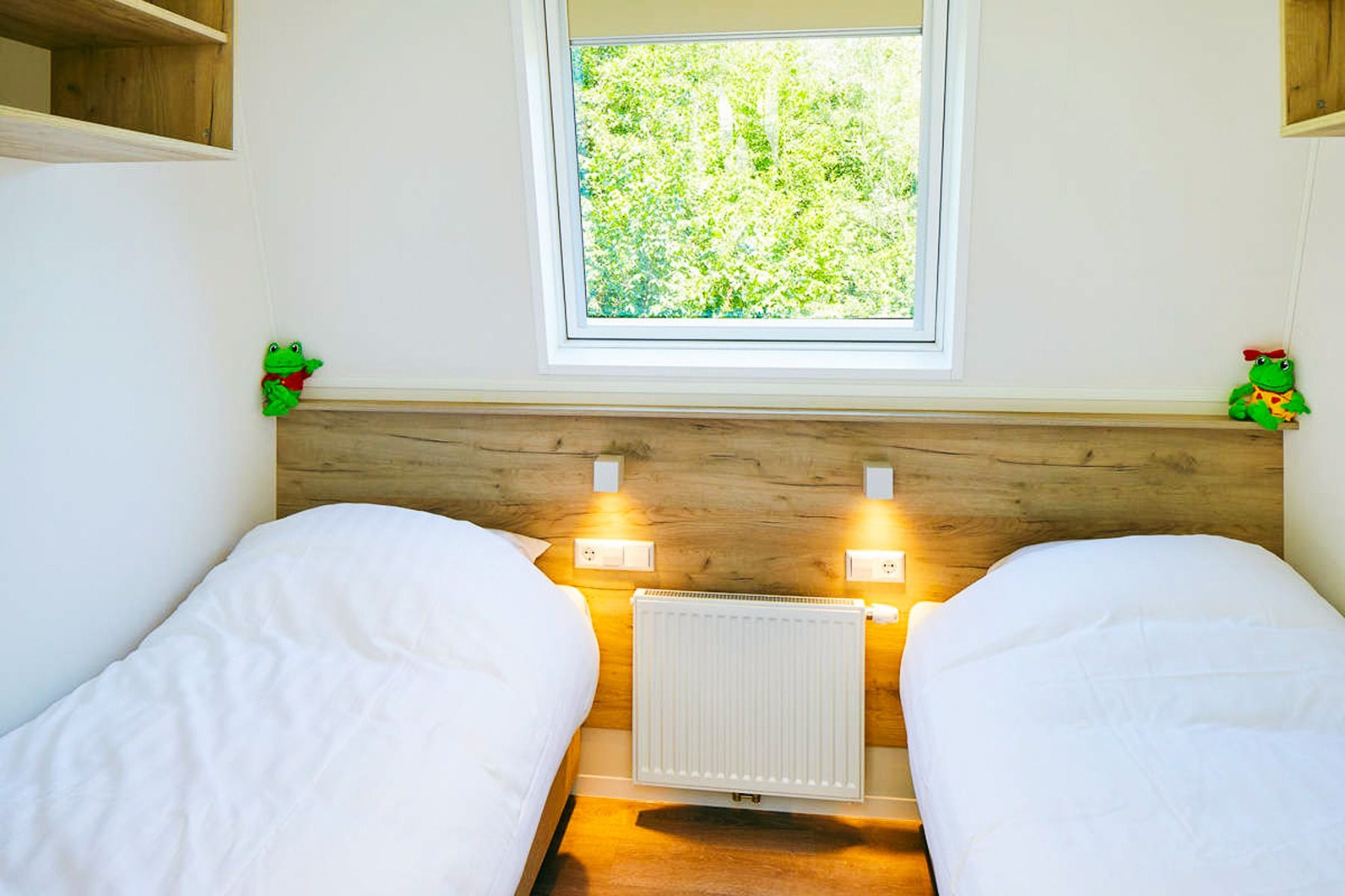 Een lichte slaapkamer op Duinrell heeft twee eenpersoonsbedden met wit beddengoed, een houten hoofdeinde, muurverlichting, een radiator, uitzicht op groene bomen door het raam en twee pluchen kikkerspeeltjes op de planken aan het hoofdeinde.