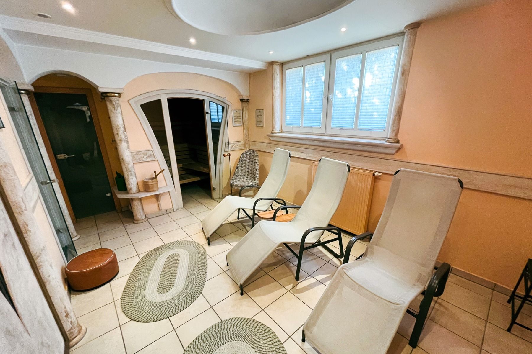 De Egger Stüberl biedt een rustige ontspanningsruimte met drie witte fauteuils, beige muren, decoratieve tapijten, boogdeuren naar een sauna en veel natuurlijk licht dat binnenvalt door de grote ramen.