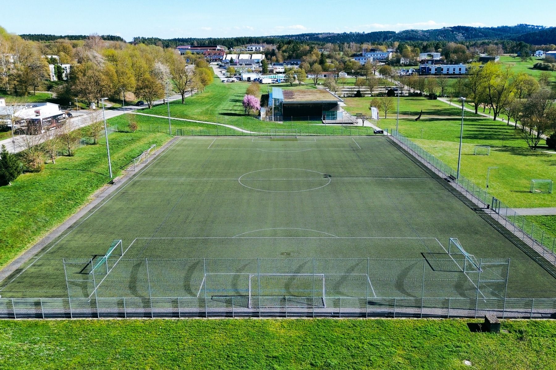 Luchtfoto van een leeg voetbalveld, ideaal voor een voetbaltrainingskamp, omgeven door groen en heuvels onder een blauwe hemel, met comfortabele accommodatie in het nabijgelegen Hotel Empfinger Hof.