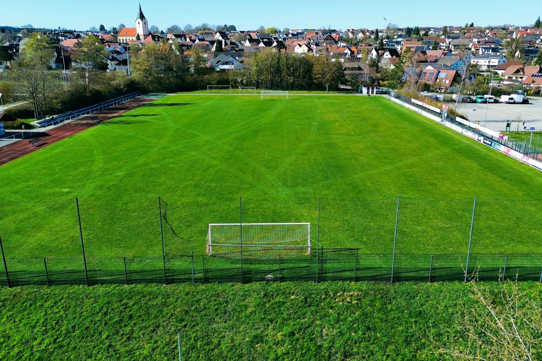 Flyfoto av en tom, grønn fotballbane med en målstolpe i forgrunnen - ideell for treningsleirer - omgitt av en løpebane, trær og hus, og med en kirke og Hotel Empfinger Hof synlig i bakgrunnen.