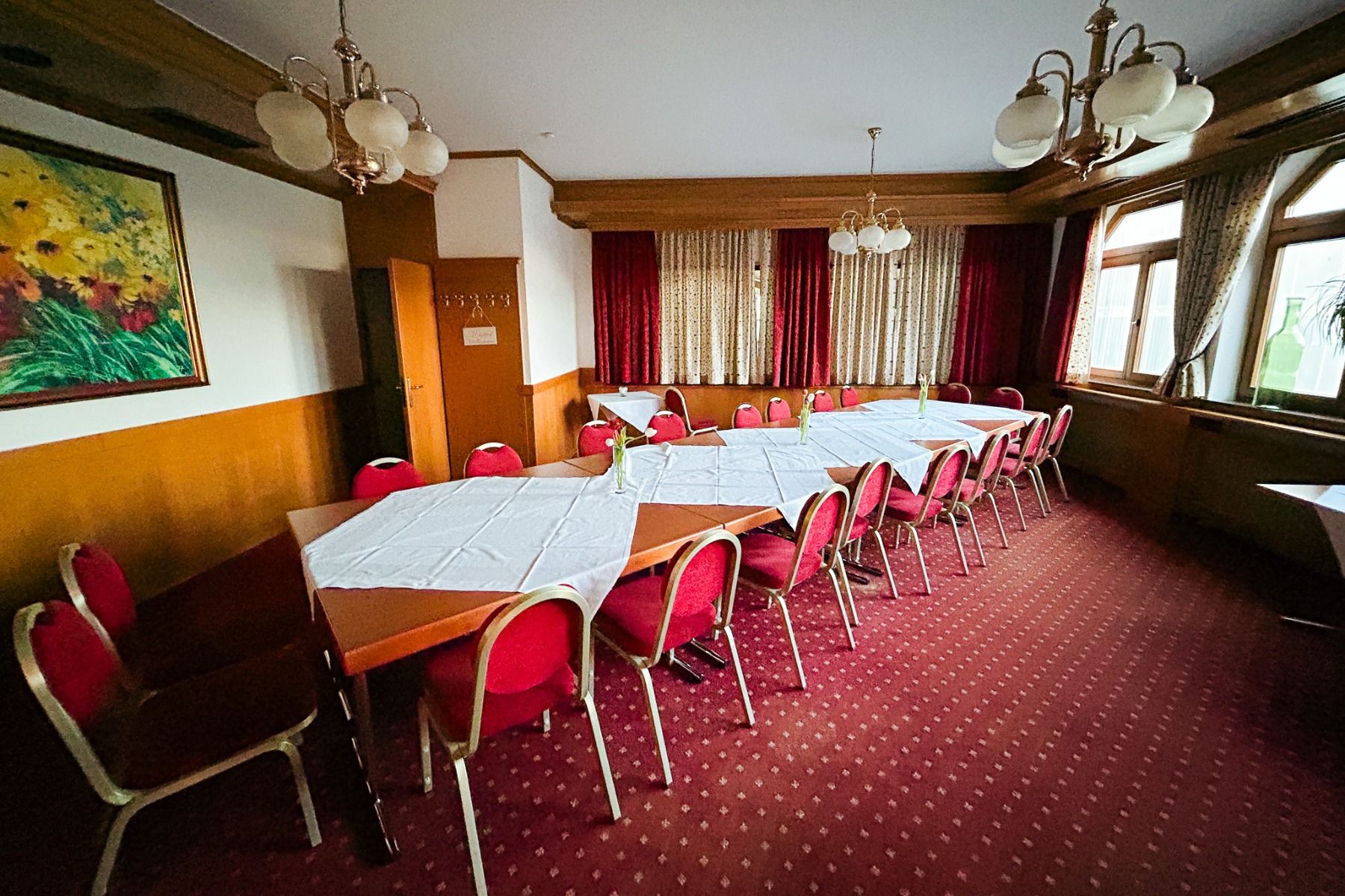Et konferanserom på Hotel Empfinger Hof har røde stoler, hvite duker, rødt teppe, trevegger, lysekroner, store vinduer med gardiner og et fargerikt maleri - ideelt for møter eller fotballtreningslager med utmerket catering.