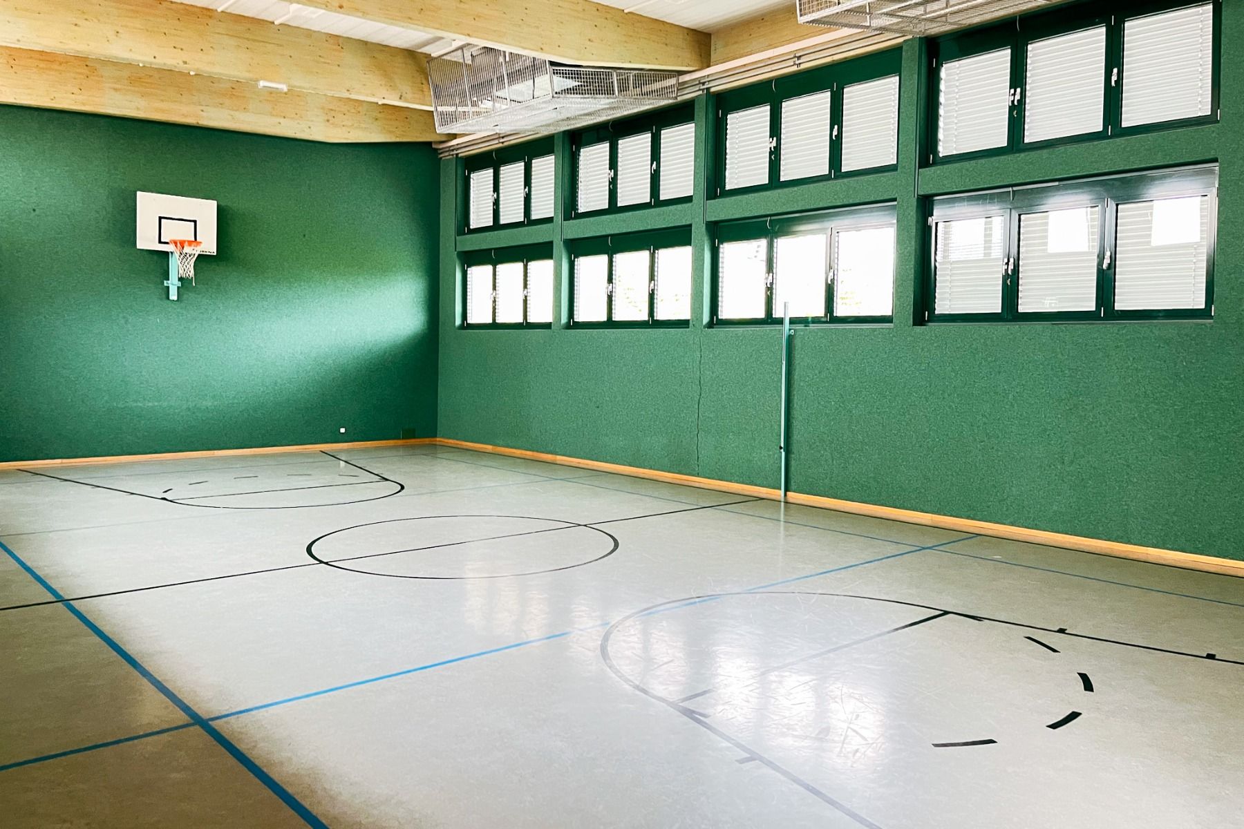 Gästehaus Burgenland Güssing heeft een fitnessruimte met een basketbalhoepel, groene muren, hoge ramen met jaloezieën en een gepolijste vloer die is gemarkeerd voor sport. Zonlicht stroomt naar binnen door de grote ramen.
