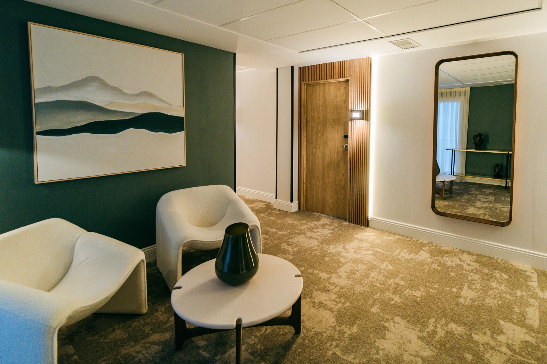 Moderne stue med to hvite lenestoler, et rundt bord med en mørk vase, et stort veggspeil og en tredør. Inspirert av Hotel Gaudea, med et landskapsmaleri på en grønn aksentvegg og myk belysning.