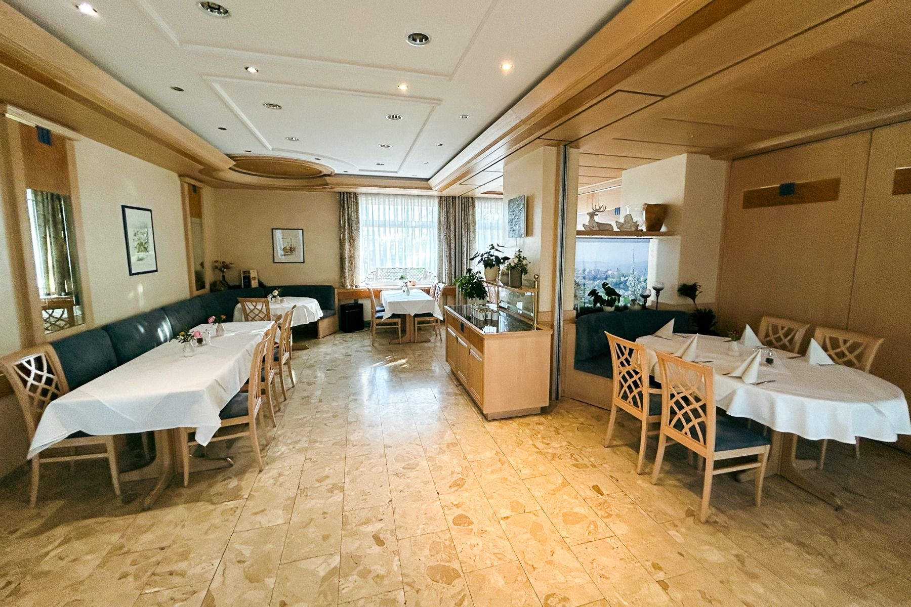Een lichte, lege eetzaal in Hotel & Restaurant Kerzenstüble verwelkomt gasten met diverse houten tafels en stoelen, witte tafelkleden, groene banken met kussens, grote ramen, weelderige planten en een lichtgekleurde tegelvloer. Deze uitnodigende sfeer vor