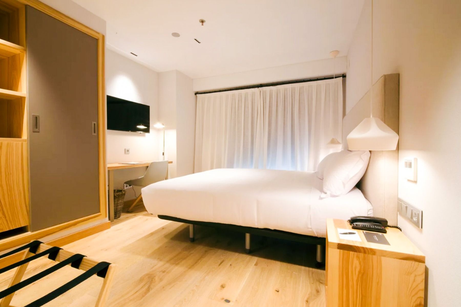 Een moderne kamer in Hotel Zenit Sevilla heeft een tweepersoonsbed, lichtgekleurd houten meubilair, een tv aan de muur, een bureau, een stoel, een groot raam met witte gordijnen en hanglampen - ideaal voor voetbalsportgroepen of zakenreizigers.