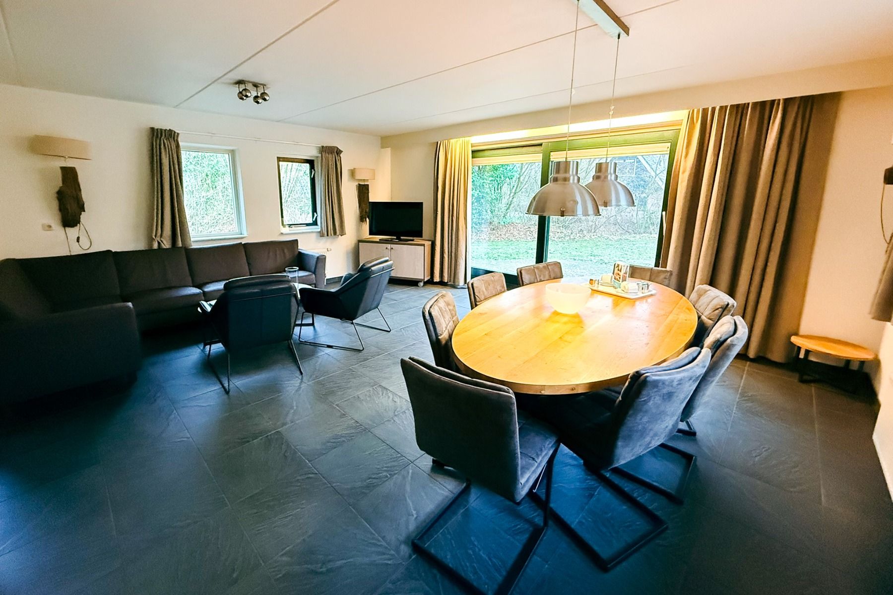 Ervaar modern wonen en dineren in Marveld Recreatie bij Groenlo, met een ronde houten tafel met grijze stoelen, donkere bank, tv, grote ramen met gordijnen met uitzicht op groen en stijlvolle donkere tegelvloeren - ideaal voor een verblijf in een bungalow