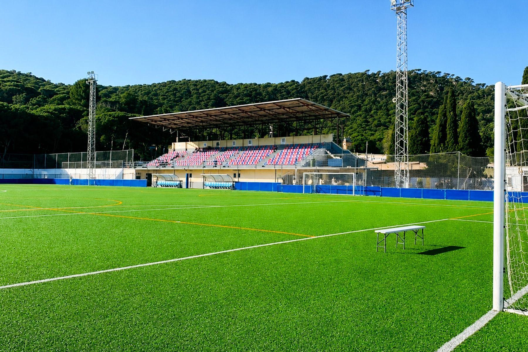 Zicht op een leeg voetbalveld met groen kunstgras bij het Van der Valk Hotel BARCAROLA aan de Costa Brava, met witte doelpalen, bank, stadionstoelen met overkapping en met bomen begroeide heuvels onder een strakblauwe hemel.