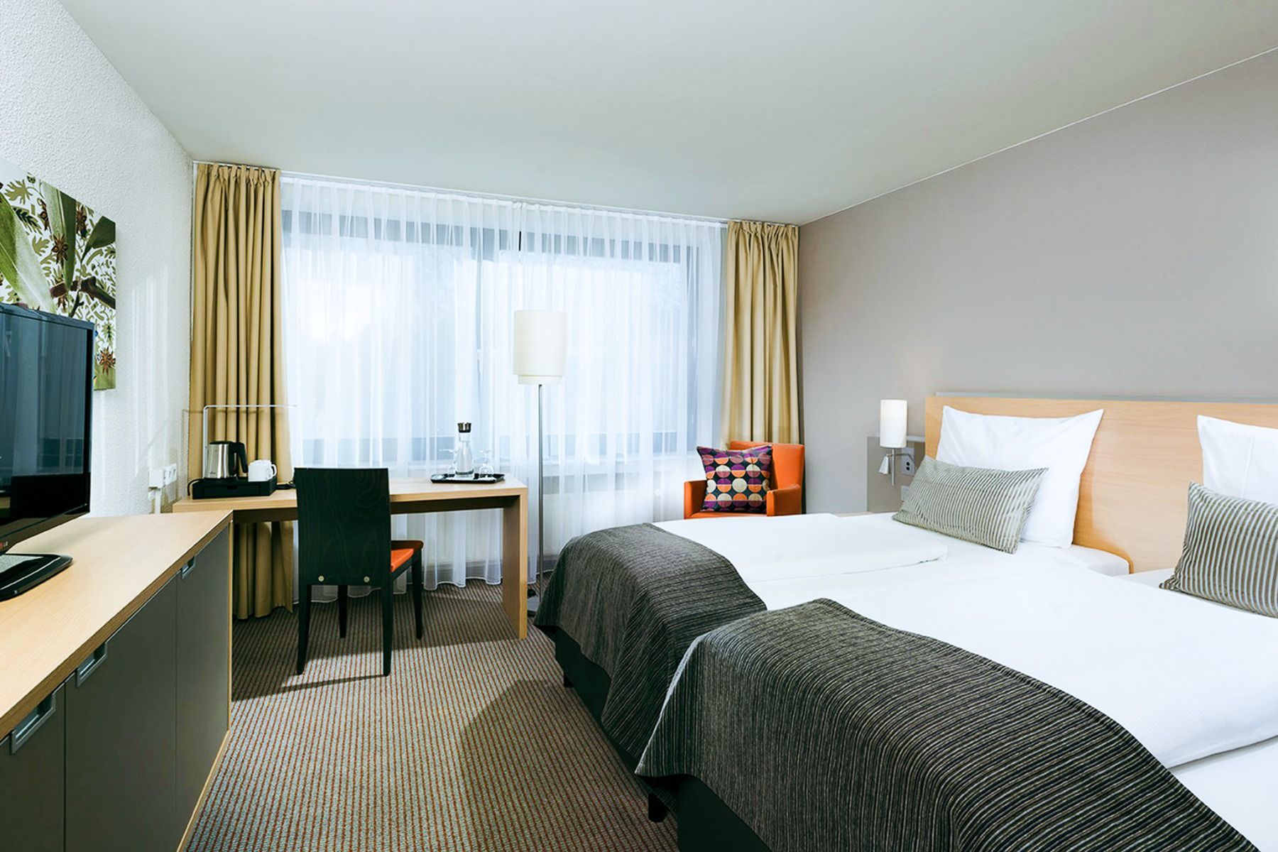 Geniet van het comfort in het Mercure Hotel in Düsseldorf, met een moderne kamer voorzien van twee keurig opgemaakte bedden, een houten bureau met stoel, een flatscreen-tv op een kast, een oranje fauteuil en een groot raam met witte vitrages en beige gord