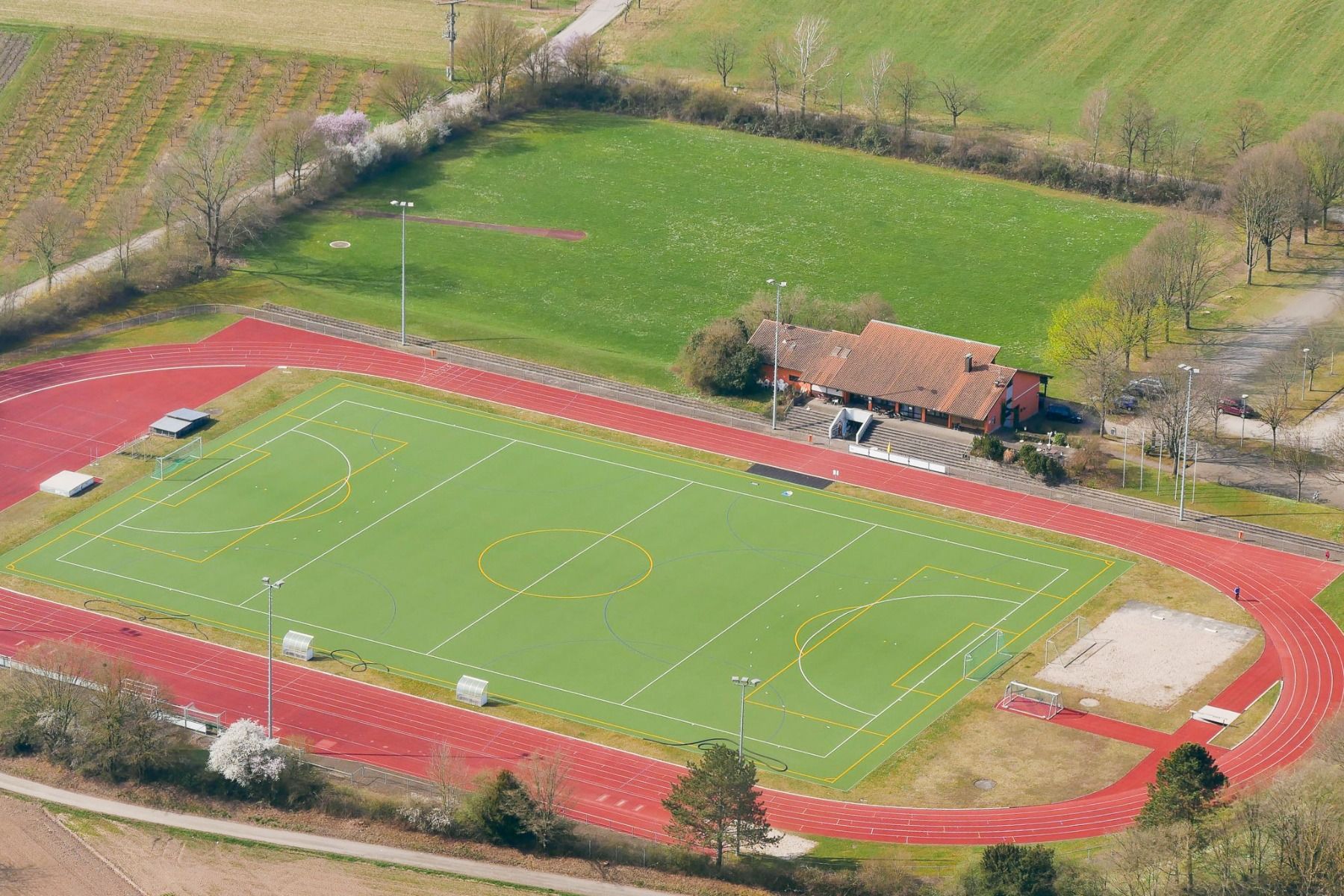 Luchtfoto van een sportcomplex met een groen voetbalveld, een rode atletiekbaan, een kleine tribune en een bruin gebouw – ideaal voor trainingskampen – omgeven door bomen en grasvelden in de buurt van het Zwarte Woud. Op het terrein bevindt zich ook het n