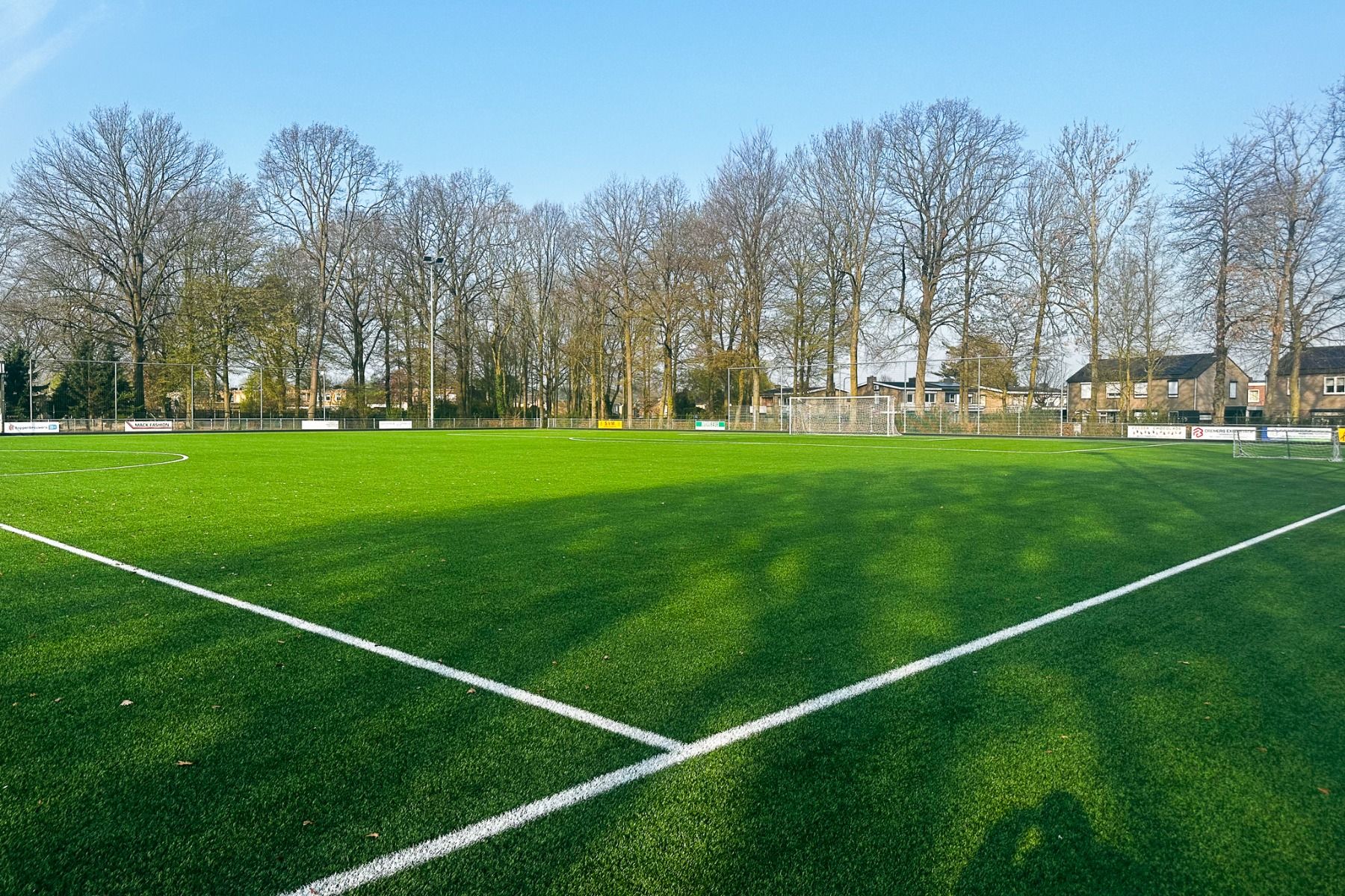 Een voetbalveld met weelderig groen kunstgras, witte lijnen en een doelpaal in het Watersley Sports & Talent Park. Achter het hek zijn kale bomen en huizen te zien en de lucht is helderblauw.