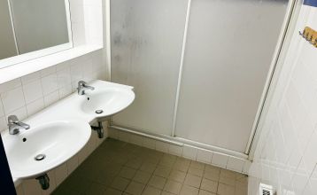 Het Gästehaus Burgenland Güssing is een groot, crèmekleurig gebouw met een rood dak en veel ramen onder een felblauwe hemel, met een aantal auto's ervoor geparkeerd en bomen in de buurt.